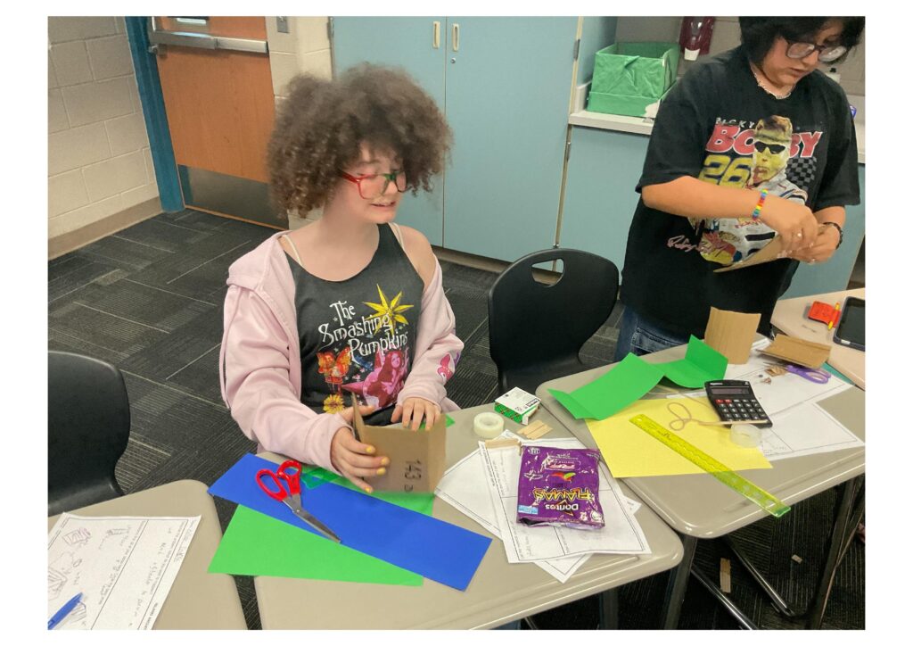 Two students work on a craft project at a table, surrounded by colorful paper, scissors, and other supplies.