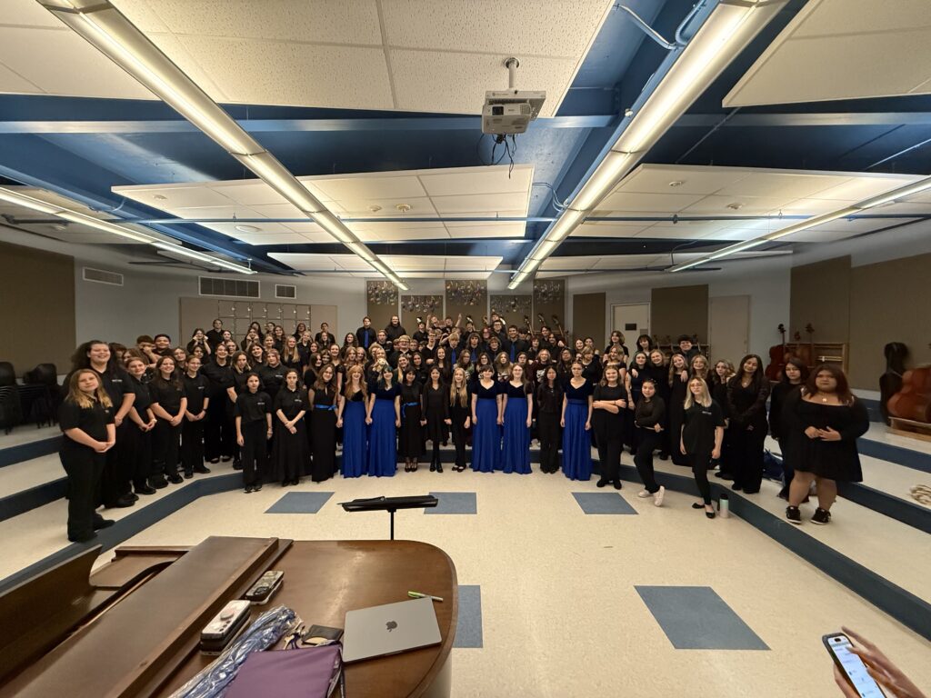 A large choir group poses together in a rehearsal space, wearing black and blue attire, with instruments in the background.