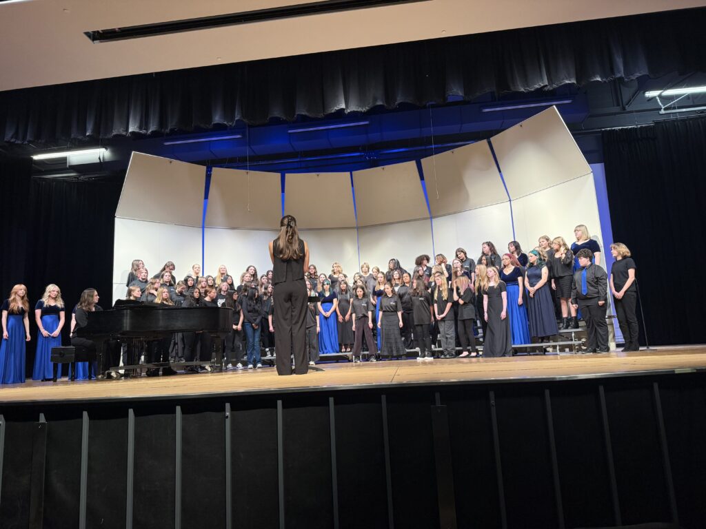 A large choir dressed in black and blue performs on stage, with a pianist accompanying them.