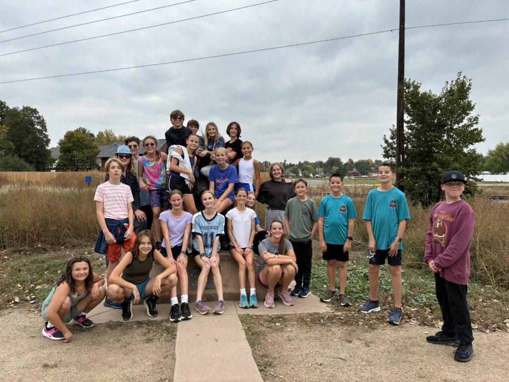 Group of children and teens posing outdoors, some sitting and others standing, with cloudy skies in the background.