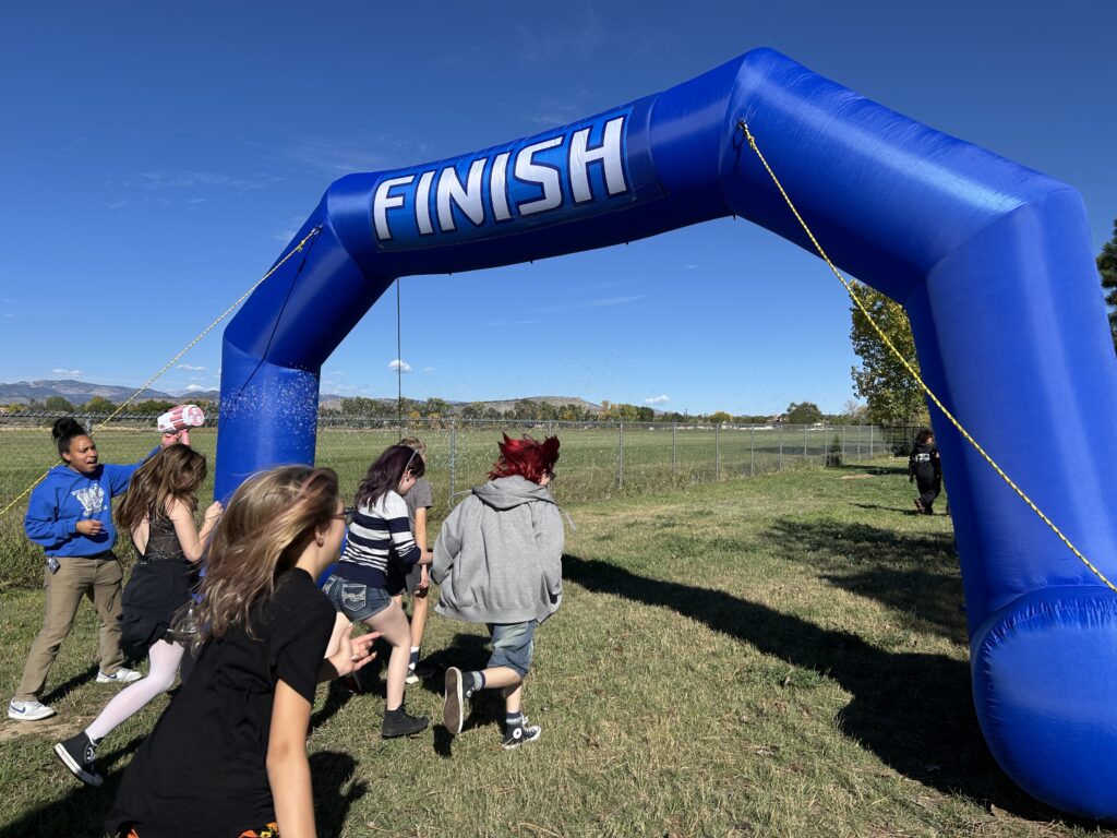 A group of children runs under a large blue inflatable finish line in a grassy field on a sunny day.