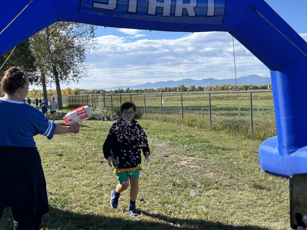 A child runs through a blue inflatable start arch while being sprayed with bubbles by a person nearby.