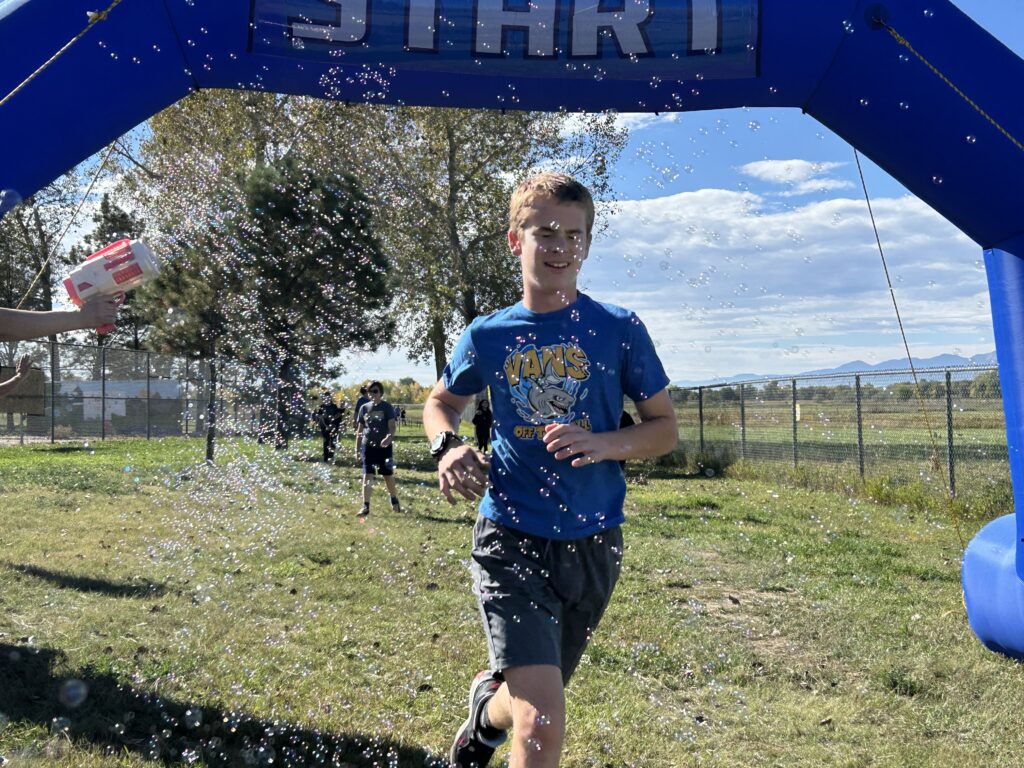 A boy runs under a blue "START" arch, surrounded by bubbles, with others in the background on a sunny day.