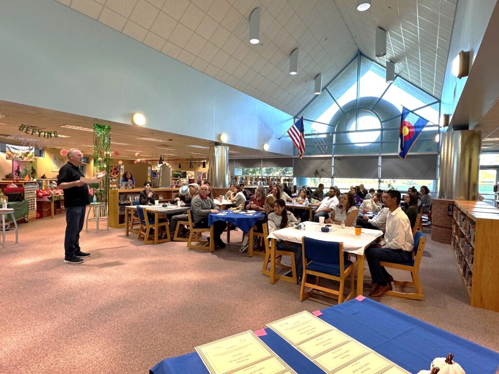 A speaker addresses an audience in a library, with tables set and flags displayed in a bright, open space.