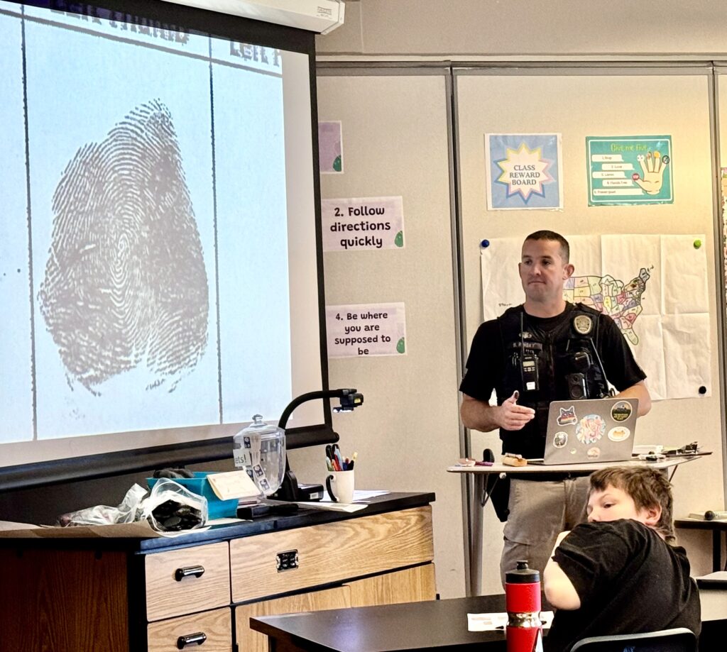 A police officer presents a fingerprint on a screen to a classroom of students, with instructions posted on the walls.