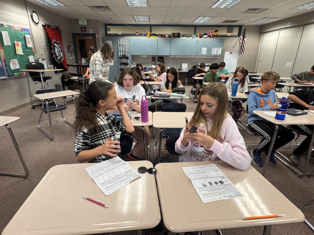 Students are seated at desks in a classroom, engaged in activities and using their phones. A teacher assists nearby.