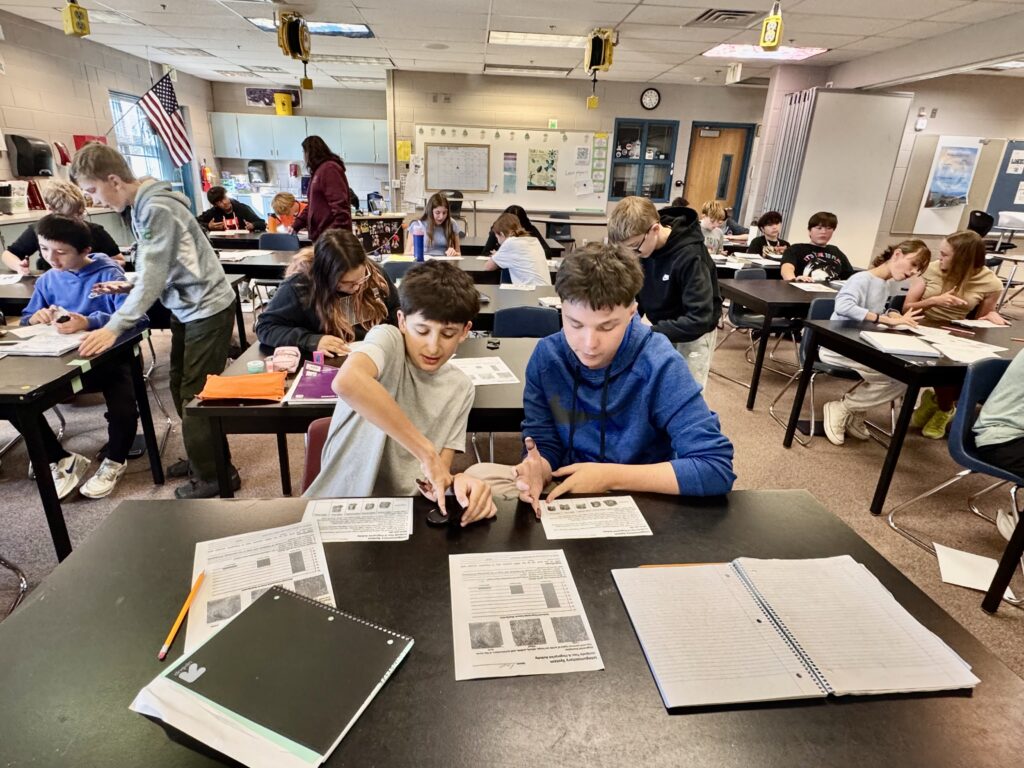 Students are engaged in a classroom activity, working at desks with papers and materials. An American flag is visible.