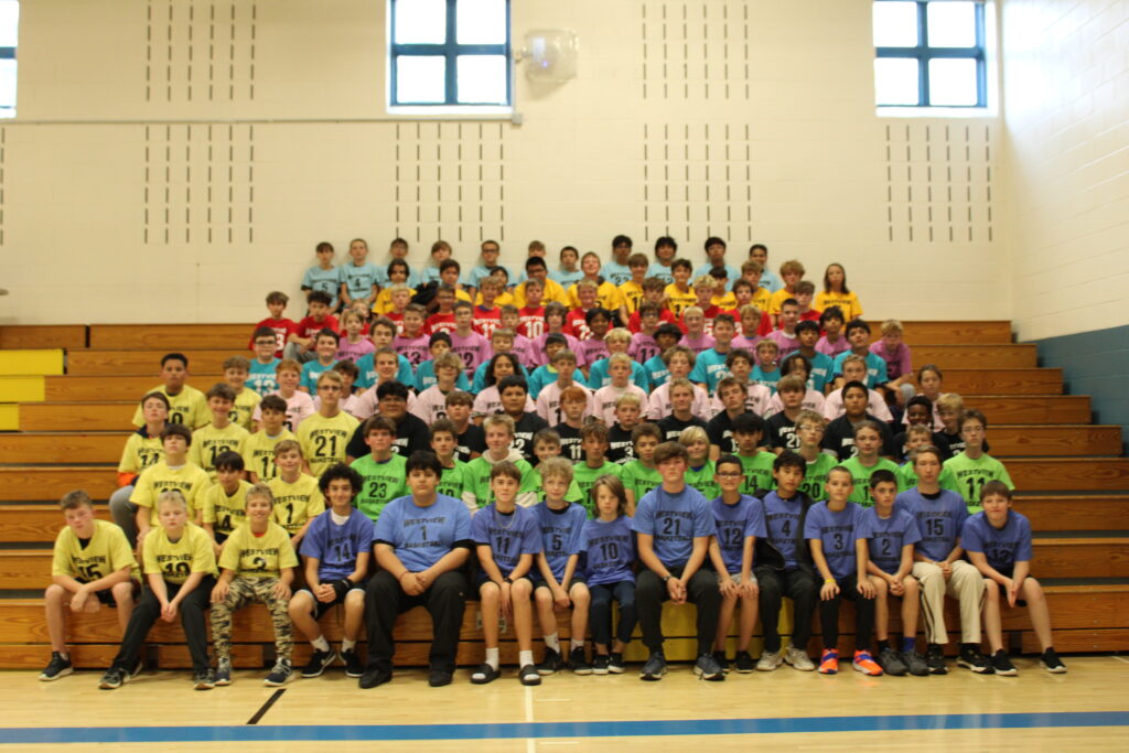 A large group of boys in colorful team shirts poses on bleachers in a gymnasium, smiling for a team photo.