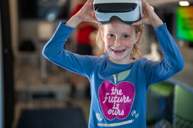 A girl with red hair smiles while holding a virtual reality headset, wearing a blue shirt with a heart design.