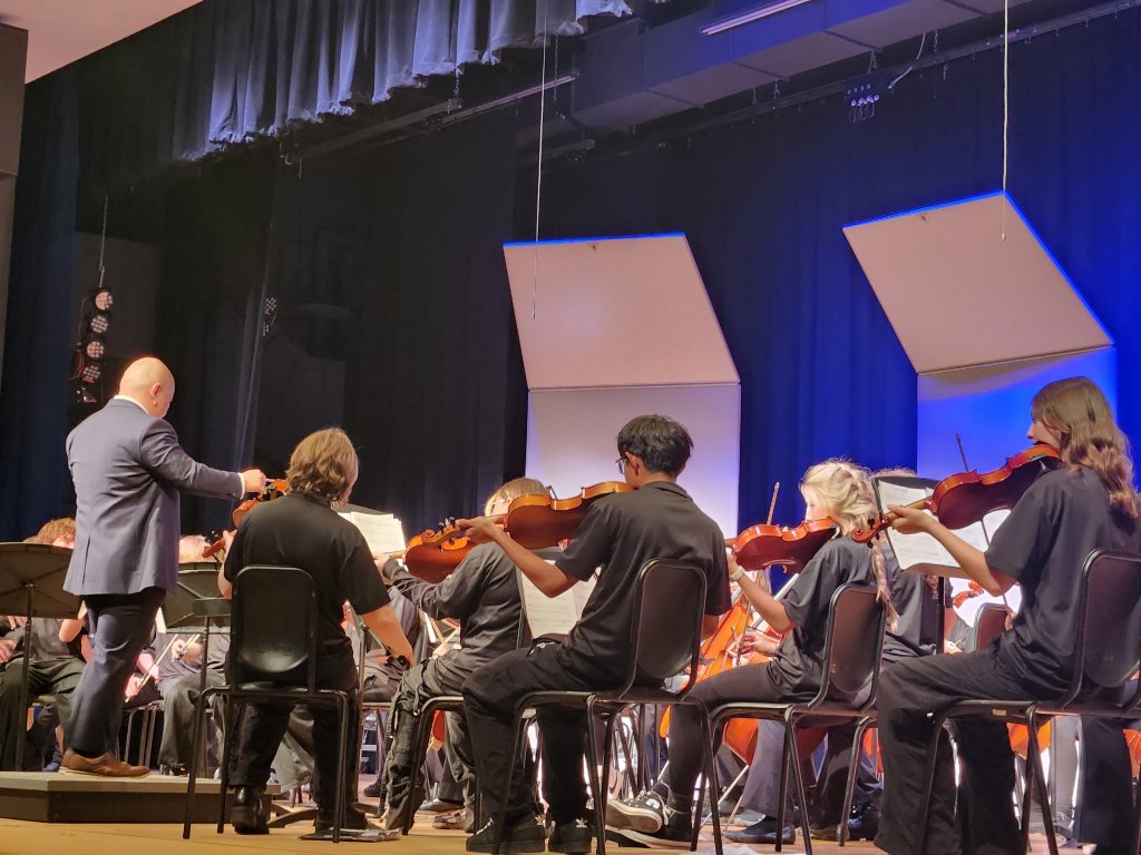 A conductor leads a youth orchestra, with musicians playing violins on stage under blue and black lighting.