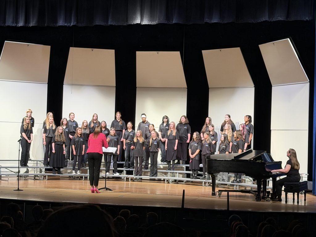 A choir of students in black attire performs on stage, led by a conductor in a red jacket, with a pianist nearby.