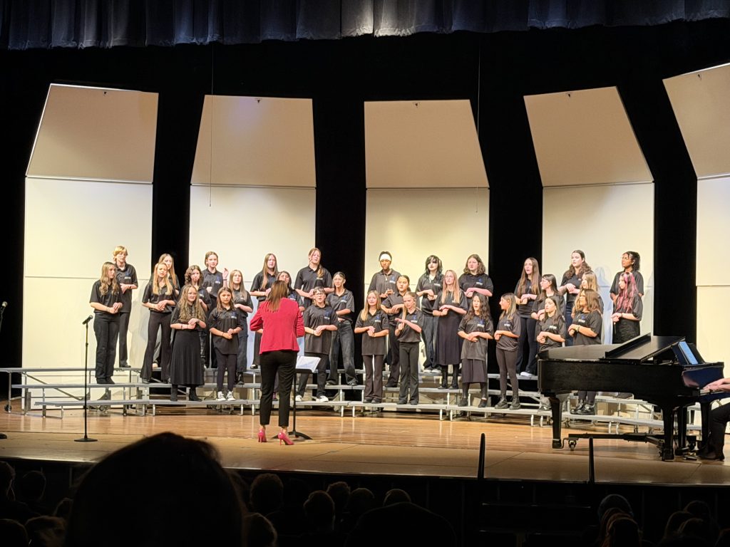 A choir of students in black attire performs on stage, directed by a conductor in a red jacket. A pianist is visible.