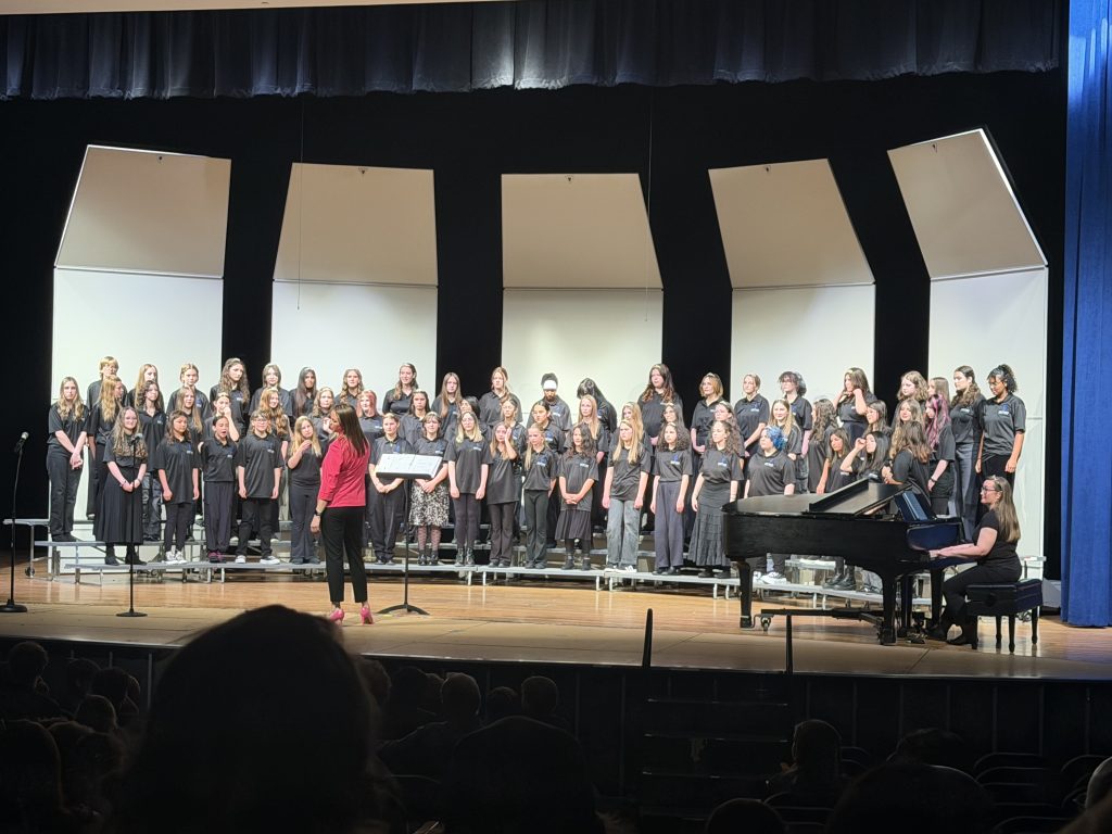 A large choir of students in black shirts performs on stage, with a conductor in a pink top and a pianist at a grand piano.