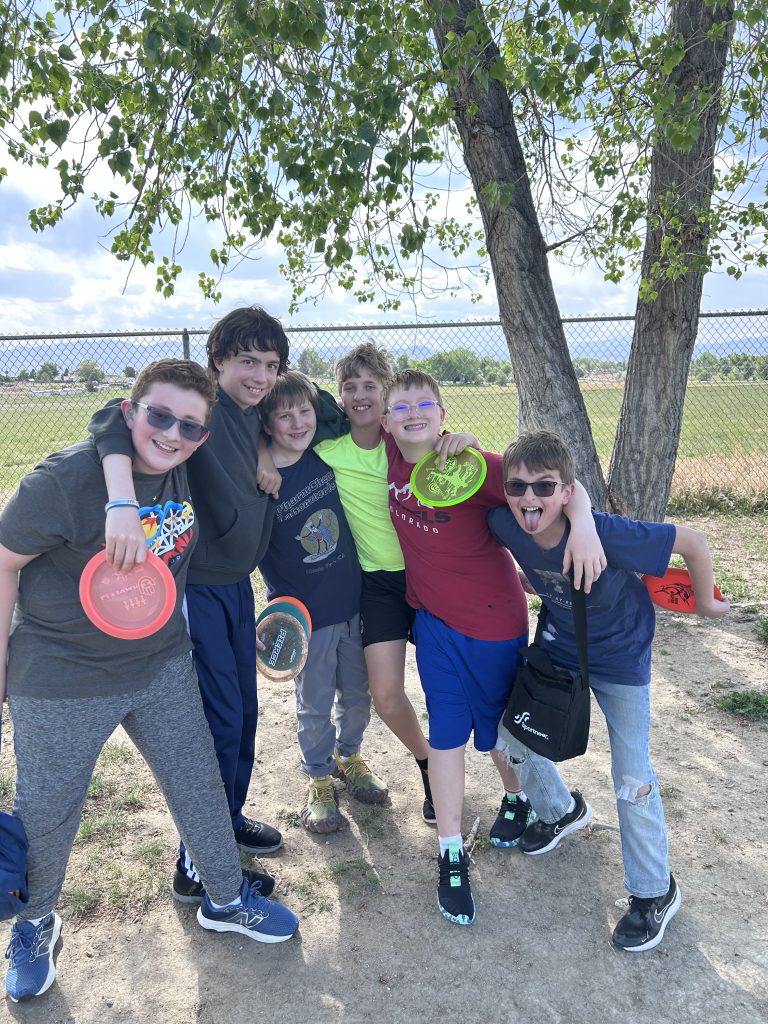 Group of five kids smiling and posing with frisbees under a tree on a sunny day, enjoying outdoor playtime.