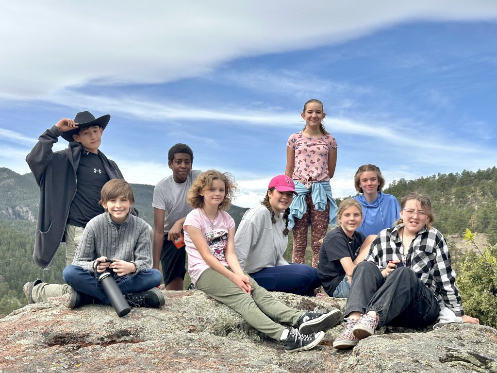A group of ten children poses on a rocky outcrop with a scenic backdrop of mountains and trees.