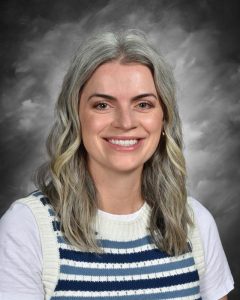 Mujer sonriente con el pelo ondulado hasta los hombros, que lleva un chaleco de punto a rayas y una camisa blanca, con un fondo gris.