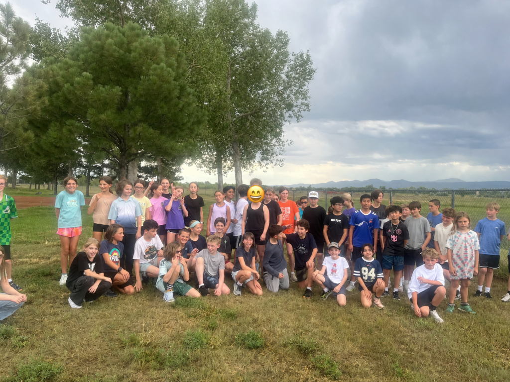 A large group of children stands outdoors, smiling together in front of trees and mountains under a cloudy sky.