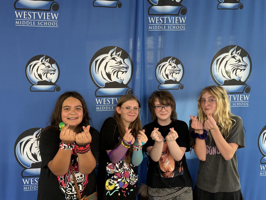 Four students pose in front of a Westview Middle School backdrop, making playful hand gestures and smiling.