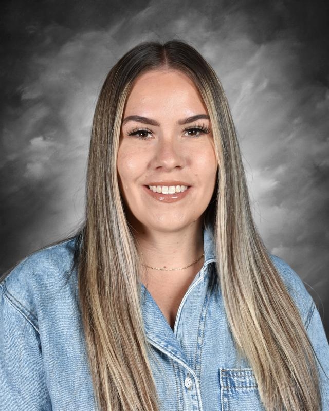 Smiling woman with long hair wearing a denim shirt against a cloudy backdrop.